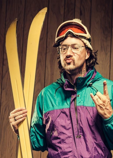 A stereotypical skier junkie with a mustache in a retro wood paneled ski lodge takes a break from the mountain slopes, his yellow skis in hand.  He wears an ugly one piece ski suit, a beanie and ski goggles.  He does a rock and roll hand sign and makes a funny face.